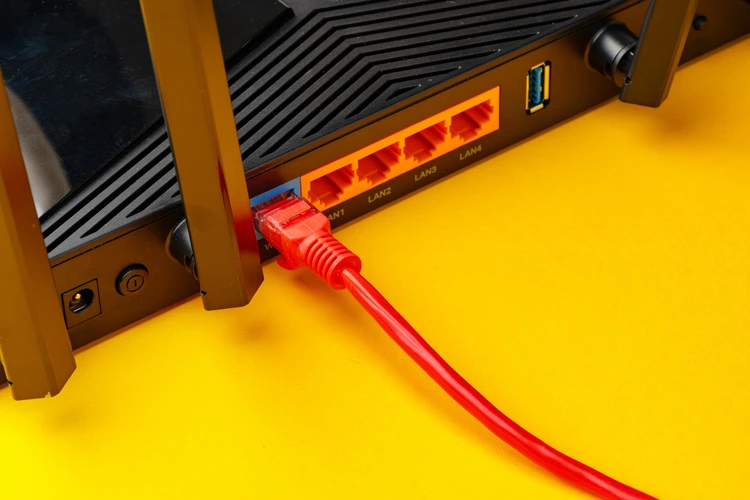 Cabo de rede vermelho conectado à porta de internet (WAN) azul na parte traseira de um roteador preto, sobre uma superfície amarela.