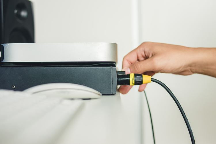 Mão de uma pessoa conectando um cabo de áudio com conector amarelo e preto em um equipamento de som em uma mesa de estúdio.